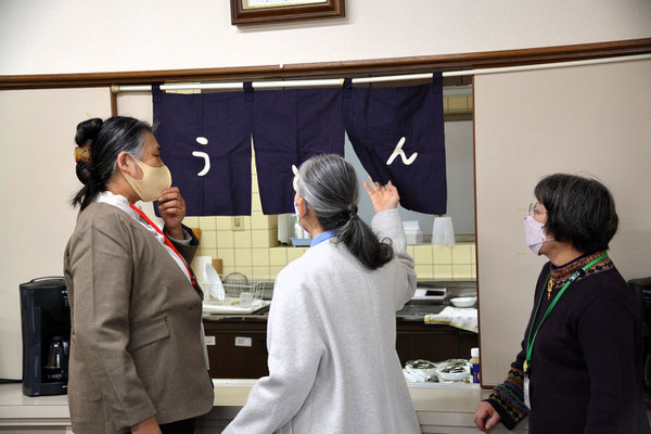 2024年12月15日（日）久しぶりに掛けられたのれんを前に、のれんの歴史などを教えて下さる光代さんの言葉に耳を傾けるお二人の図でせ。