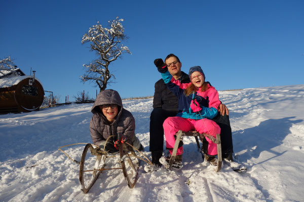 Ein Bub liegt auf seiner Rodel, Vater und Tochter sitzen auf einer zweiten Rodel daneben, lachen und rodeln den Schneehang hinunter.