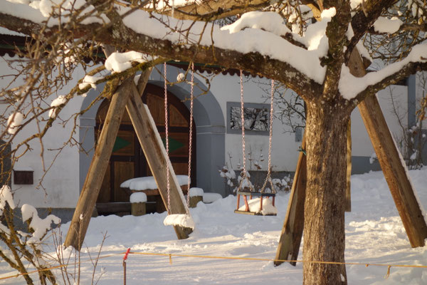Im Hintergrund ist der verschneite Bauernhof zu sehen mit seinem Hoftor, im Vordergrund die verschneite Schaukel.