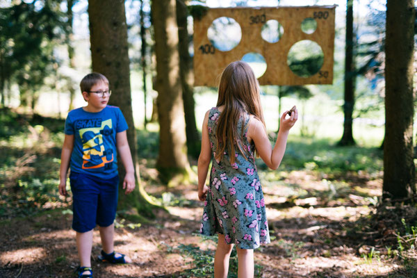 Ein Bub und ein Mädchen werfen im Wald Zapfen durch ein Brett mit Löchern, das zwischen den Bäumen hängt.