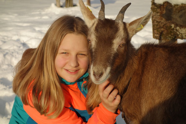 Mädchen mit langem blonden Haaren lacht und kuschelt mit einer kleinen braunen Ziege. Im Hintergrund liegt Schnee.