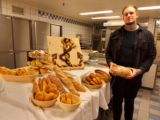 Absolvent steht mit einem Brot in der Hand vor seinen Gebäckstücken in der Backstube