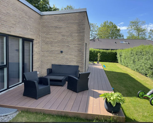 Moderne Terrasse mit Bambus-Terrassendielen Holguin in warmem Braunton, ausgestattet mit Markisen, Loungemöbeln und Topfpflanzen vor einem Flachdachhaus aus Klinker bei Sonnenschein.