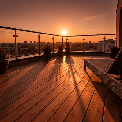Moderne Dachterrasse mit Holzdielen, Glasgeländer, Sitzmöbeln und Kübelpflanzen im warmen Licht des Sonnenuntergangs.