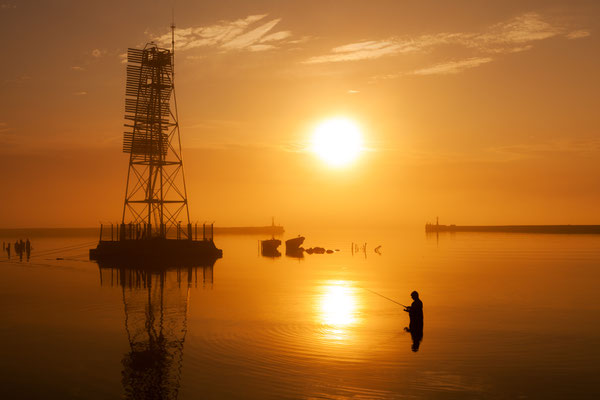 Fisherman in the water near lighthouse on the outstanding sunset background with reflection on foggy weather on Baltic sea - Copyright Konstantin Romanov