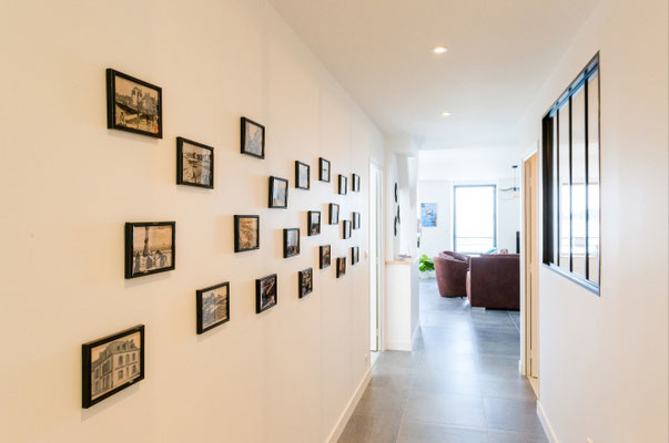 Couloir d’appartement avec décoration murale encadrée, sol en carrelage gris et perspective vers la pièce de vie lumineuse
