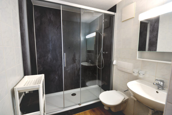 Vom Badezimmereingang Blick auf die Dusche, die sich über die ganze Wandbreite zieht,, mit duchsichtiger Dusch-Schiebetür, Duschwand hinten schiefergrau, rechts ander Wand Tooilette und Waschbecken mit Spiegelschrank