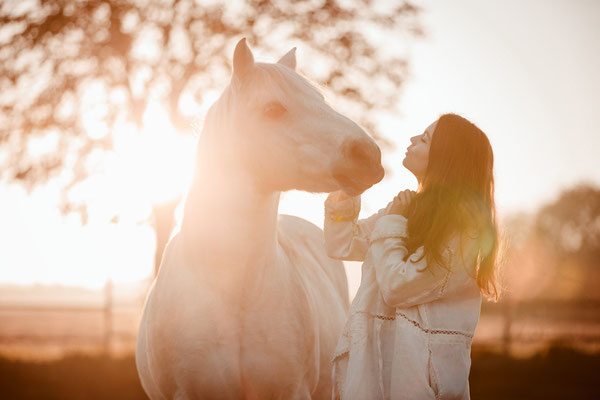 Eine Frau in einem Kleid steht mit einem weißen Pony im morgendlichen Gegenlicht.