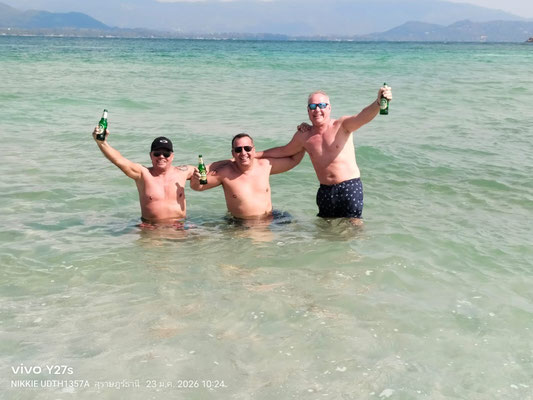 Drei Männer stehen lachend mit Bierflaschen im türkisblauen, flachen Wasser von Koh Samui.