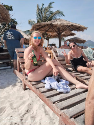 Eine Frau entspannt mit einem kühlen Getränk und Sonnenbrille auf einer Holzliege am Strand.