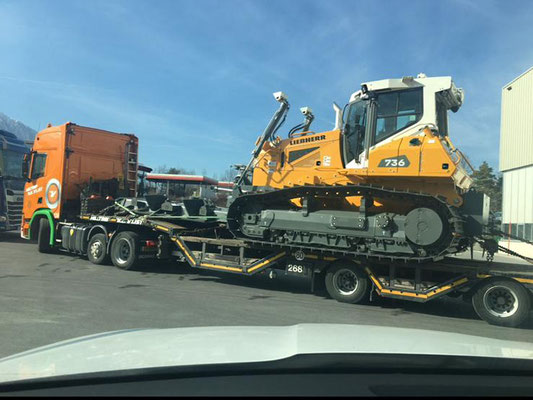 Orangefarbener Schwerlastzug, der einen gelben Kettenbagger auf einem Tieflader-Auflieger auf einer Bergstraße transportiert.
