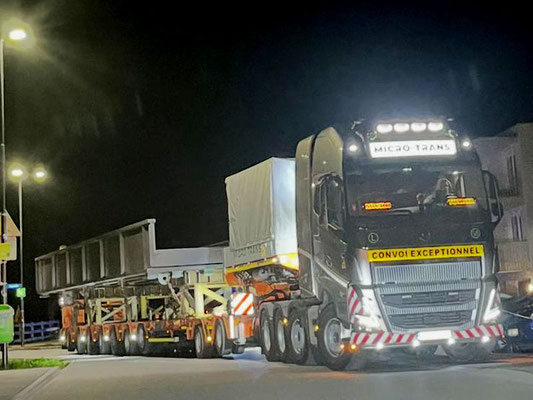 Silberner LKW transportiert nachts einen riesigen weißen Turmabschnitt oder Behälter auf einem Tieflader, beleuchtet von Straßenlaternen.