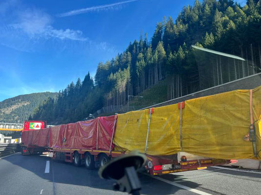 Rückansicht eines Schwerlasttransporters mit verdeckter Ladung, der mit Warnleuchten auf einer Straße in einer teilweise verschneiten, bewaldeten Landschaft fährt.