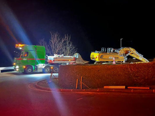 Weißes Begleitfahrzeug mit Warnleuchten vor einem roten Schwerlasttransporter, der einen Kasten-Container transportiert.