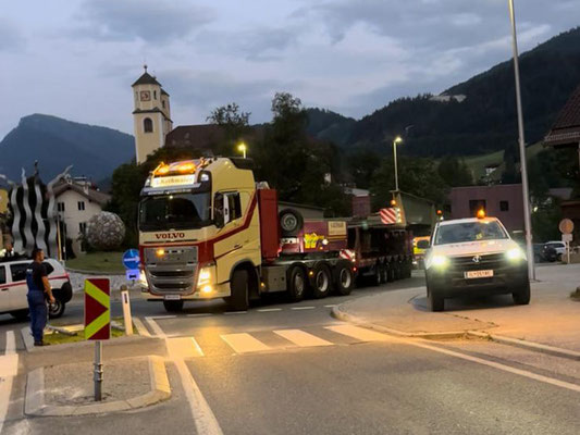 Weißer LKW und ein weißes Begleitfahrzeug mit Warnblinkern davor, die eine verpackte Last auf einer Bergstraße transportieren.