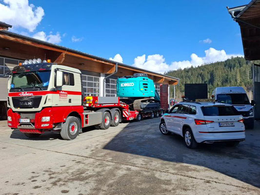 Roter LKW und ein weißes Begleitfahrzeug davor, die eine verhüllte Ladung auf einer Straße in den Bergen transportieren.