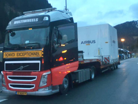 Grauer LKW mit roter Front, der einen weißen Kasten-Container auf einem Tieflader bei Nacht in einem Industriegebiet transportiert.