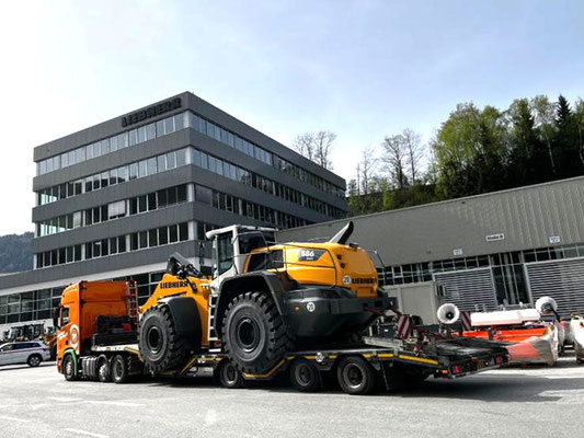 Gelber Kettenbagger auf einem Tieflader-Auflieger, geparkt vor einem modernen Bürogebäude.
