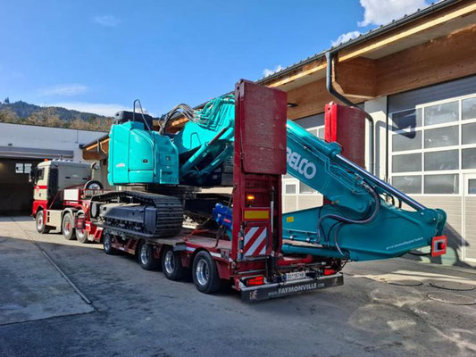 Türkisfarbener LKW zieht einen Tieflader mit einer riesigen blauen Turbinen- oder Maschinenkomponente, geparkt in einem Hof vor einer Ladehalle und Bergen.