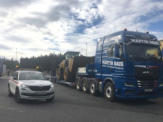 Blauer Schwerlastzug mit einem kleinen Begleitfahrzeug davor, die sich auf einer Straße in der Nähe von bewaldeten Hügeln fortbewegen.