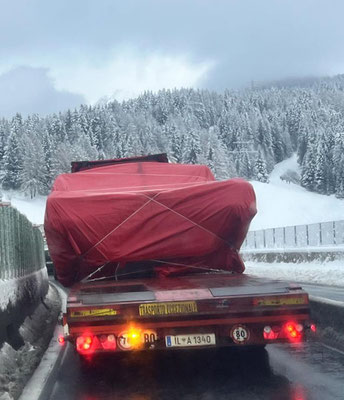 Rückansicht eines Schwerlasttransporters mit verhüllter, roter Ladung, der mit Warnleuchten auf einer nassen Straße in einer verschneiten Tannenwaldlandschaft fährt.
