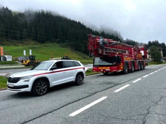Weißes Begleitfahrzeug mit Warnleuchten vor einem roten Schwerlasttransporter, der eine große, verpackte rote Industriekomponente auf einer Bergstraße transportiert.