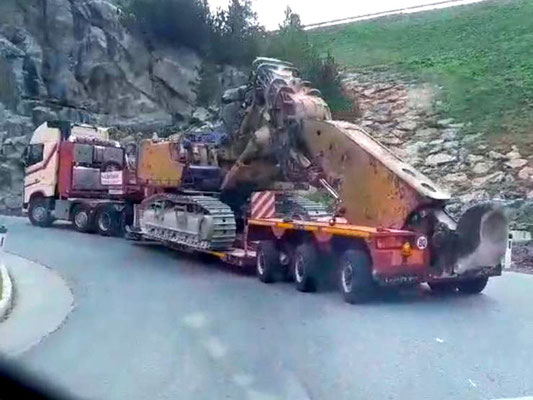 Blauer Schwerlastzug mit einem kleinen Begleitfahrzeug davor auf einer kurvigen Bergstraße, die eine verpackte Ladung transportieren.