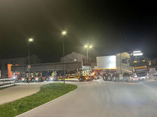 Roter Schwerlastzug und ein weißes Begleitfahrzeug davor, die nachts eine verpackte Last auf einer Straße transportieren, beleuchtet von Straßenlaternen.