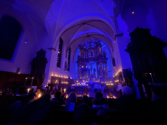 Hochaltar, blau beleuchtet, mit Kerzenhaltenden Menschen im Vordergrund