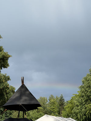 Ein Regenbogen über einem Zeltdach einer Jurte zwischen Bäumen.