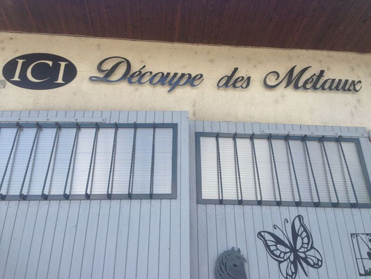 Enseigne d’atelier avec lettrage métal découpé « ICI Découpe des Métaux » au-dessus de grandes portes métalliques, illustrant la découpe des métaux et la découpe des matériau, avec un motif de papillon.