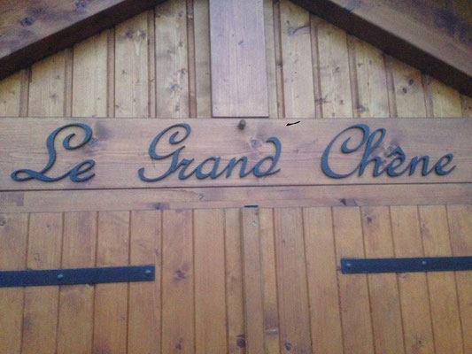 Lettre métal découpée noire formant l’inscription « Le Grand Chêne » fixée sur des portes en bois, illustrant la découpe des métaux pour la personnalisation de bâtiments.