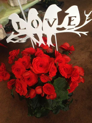 Lettre métal découpée blanche formant le mot « LOVE » avec des oiseaux, placée au-dessus d’un bouquet de fleurs rouges, exemple de découpe des matériau décoratif.