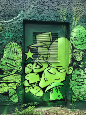Grüne Wandgestaltung im Comic-Stil auf einem Verteilerkasten. Das moderne Charakter-Design ist ein Beispiel für urbane Kunst am Bau, verfügbar als Dienstleistung bundesweit.