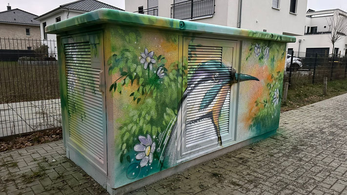 Künstlerisches Graffiti-Mural auf einem Trafohaus, das einen großen, farbenfrohen Eisvogel und weiße Blüten vor einem grün-gelben Hintergrund zeigt. Professionelle Fassadengestaltung von Appolloart zur ästhetischen Aufwertung öffentlicher Räume und als Va