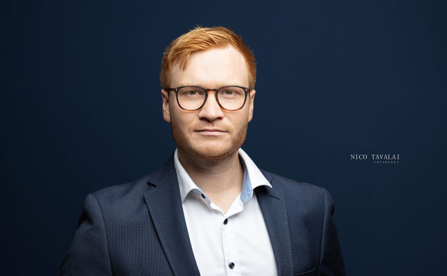 Bewerbungsfoto eines Mannes mit Brille im dunkelblauen Sakko vor neutralem Hintergrund