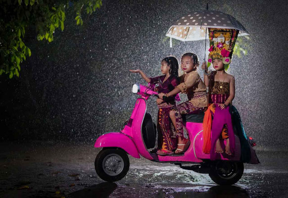 Three young girls on a Vespa scooter, dressed in dance attire, riding through a light rain.
