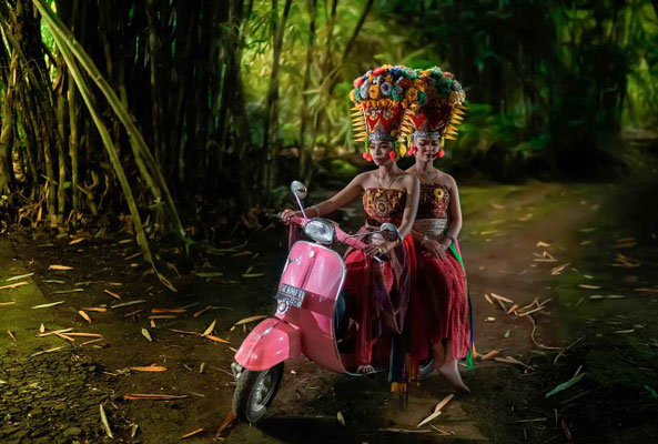 Two girls in dance attire riding a Vespa through a peaceful bamboo forest.