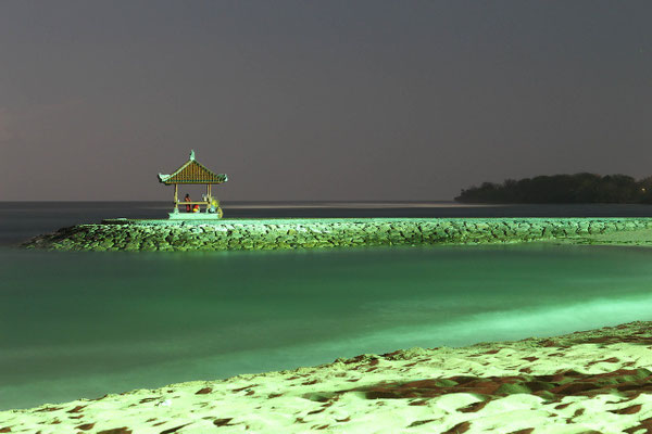 A moonlit pavilion in Sanur glowing over a calm shoreline at night.