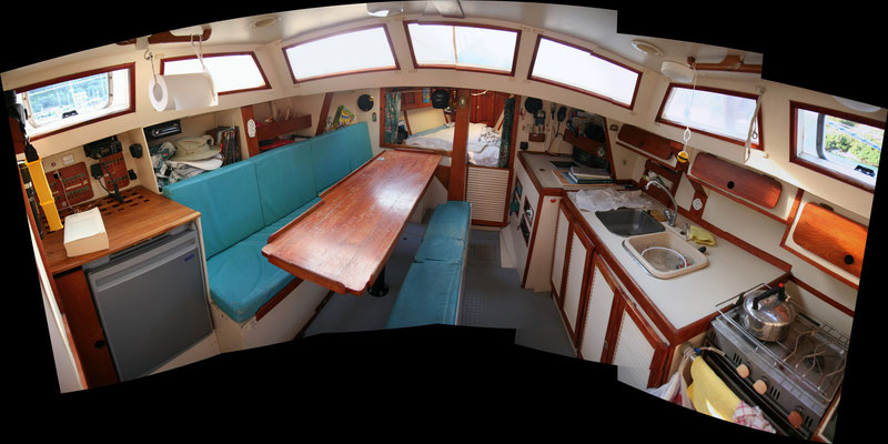 (2009) Photo panoramique prise de la descente : de bâbord à tribord, le frigo, avec au-dessus la VHF et le tableau électrique, puis le carré (transformable en couchette double), la couchette double avant, la table à cartes, la cuisine .