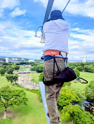 関西メンズマッサージサロン「forest hands」セラピストが万博記念公園の巨大アスレチック「万博BEAST」の空中一本橋を渡る後ろ姿。背景には広大な緑の景色と遠くに見える太陽の塔。