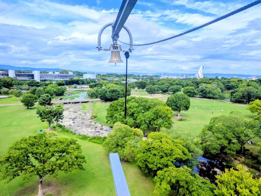 関西メンズマッサージサロン「forest hands（フォレストハンズ）」のブログ用写真。万博記念公園の巨大アスレチック「万博BEAST」の空中一本橋の先端に吊るされた鐘と、眼下に広がる広大な芝生や緑の絶景。