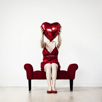 Manuela Deigert Projekte Selbstportrait auf einem kleinen roten Sofa sitzend und einen roten Herzluftballon vor mein Gesicht haltend