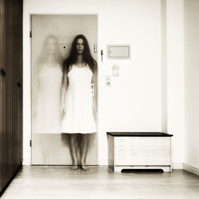 Manuela Deigert Projekte unscharf doppeltes Selbstportrait als Langzeitbelichtung in einem weißem Kleid vor einer Tür in einem Zimmer stehend