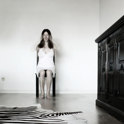 Manuela Deigert Projekte Selbstportrait als Langzeitbelichtung mit einem weißen Kleid alleine auf einem Stuhl in einem Zimmer sitzend