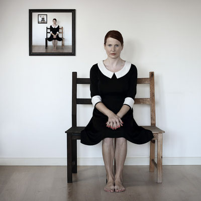 Manuela Deigert Fotokunst Start, Frame in Frame, Selbstportrait in einem schwarzen Kleid auf einem Stuhl sitzend, an der Wand ein Rahmen mit demselben Abbild 