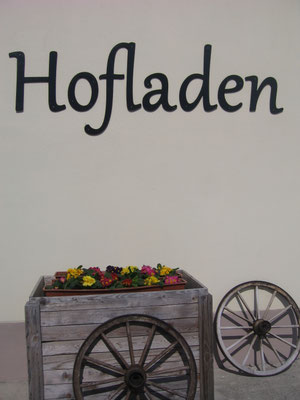 Die Aufschrift "Hofladen" mit einer Blumenkiste davor und zwei Holzrädern