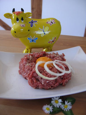Gehacktes mit einem Ei und Zwiebelringen. Dahinter eine gelbe Kuhfigur mit Schmetterling-Stickern