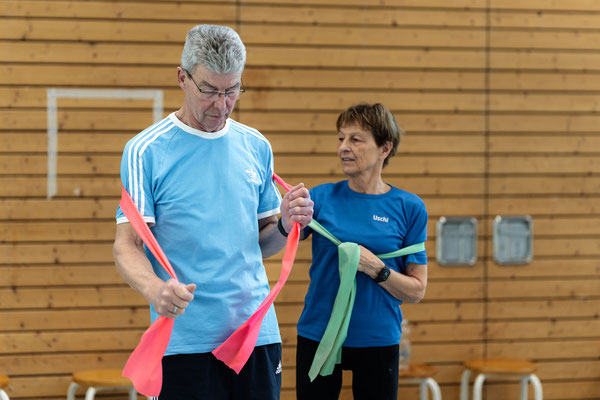 Übungsleiterin begleitet einen Teilnehmer der Herz- und Rehasportgruppe bei einer Übung mit Fitnessband in der Sporthalle.
