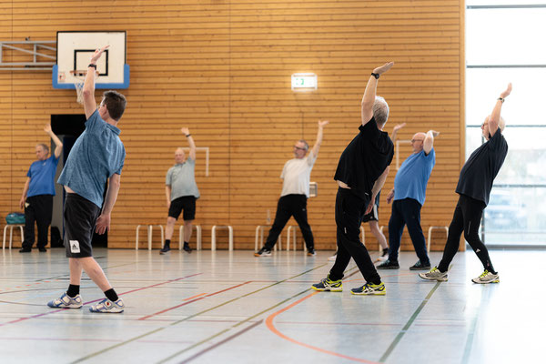 Teilnehmer der Herz- und Rehasportgruppe führen gemeinsam koordinierte Dehn- und Bewegungsübungen in der Sporthalle durch.